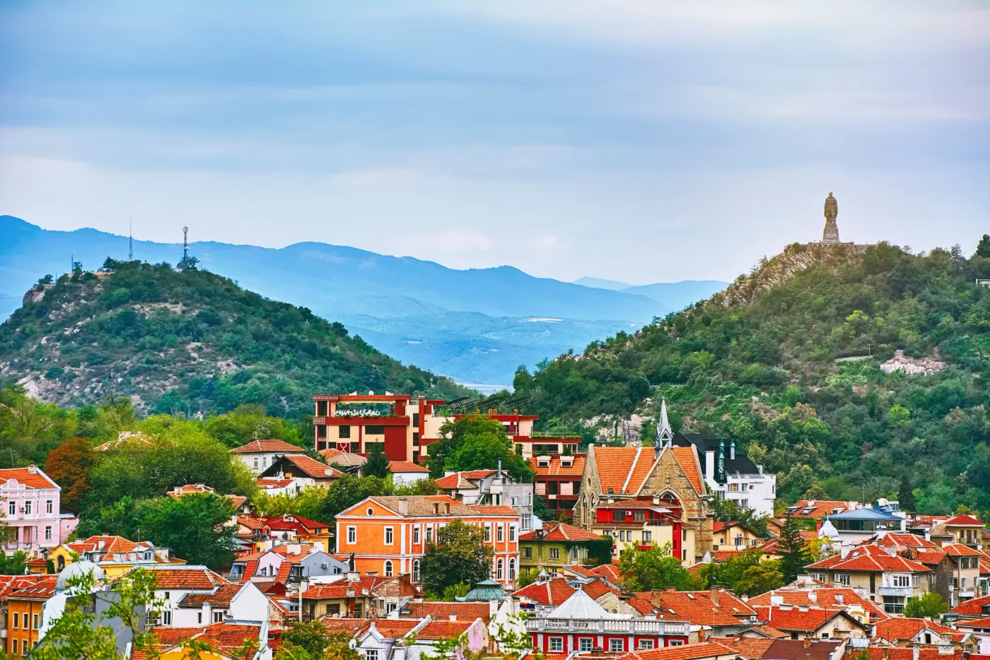 Letecký pohled na Plovdiv s historickými budovami, zelenými pahorky a památníkem Alyoša na vrcholu kopce.